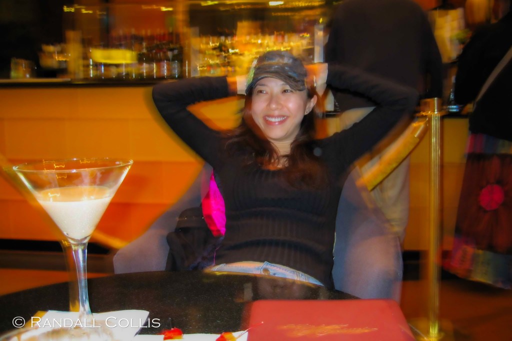 A woman relaxes in a cozy setting, smiling as she leans back with her arms behind her head, in front of a table featuring a large martini glass and a small plate of food.