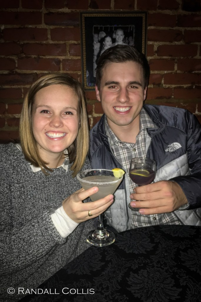 A smiling couple holds drinks at a table with a brick wall backdrop, capturing a joyful moment together.