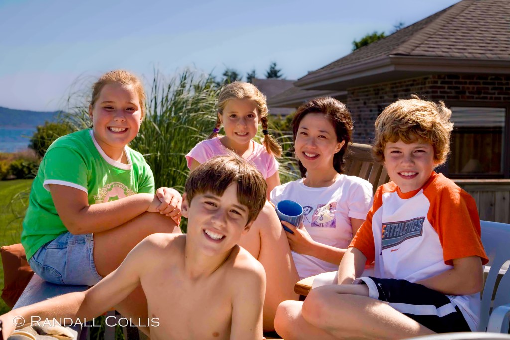 A group of five children, including three girls and two boys, smile while sitting together outdoors. They are surrounded by green grass and a house in the background, with a serene view of a lake or ocean in the distance.