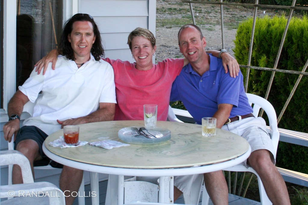 Three friends sitting together at a table outdoors, enjoying drinks and smiling at the camera.